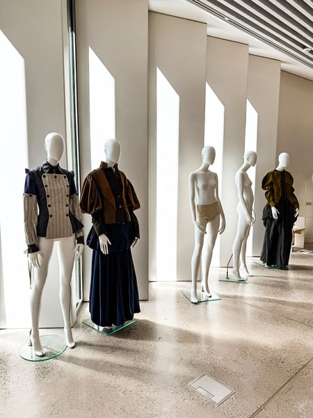 Eine Reihe weißer Schaufensterpuppen präsentiert verschiedene avantgardistische Modeentwürfe in einem hellen Ausstellungsraum der LCI Barcelona. Das Sonnenlicht fällt durch hohe, schmale Fensteröffnungen und erzeugt ein Spiel aus Licht und Schatten auf den Figuren und dem polierten Betonboden.