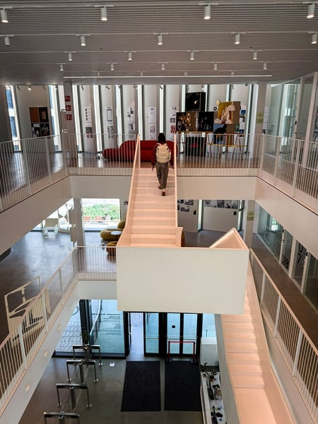 Blick von oben in das lichtdurchflutete Atrium der LCI Barcelona. Eine zentrale, weiße Treppe führt in die oberen Stockwerke, wo Ausstellungsbereiche mit dem markanten roten Sofa und Kunstobjekten zu sehen sind. Die Architektur ist durch klare Linien, weiße Geländer und eine Lamellendecke geprägt.
