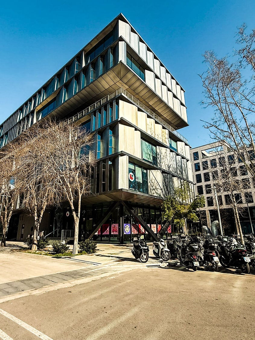 Eine Eckansicht des modernen LCI Barcelona Campus-Gebäudes mit seiner charakteristischen, verschachtelten Architektur und Aluminium-Fassade. Vor dem Gebäude an der Straßenecke stehen mehrere geparkte Motorroller unter kahlen Bäumen bei hellem Tageslicht.