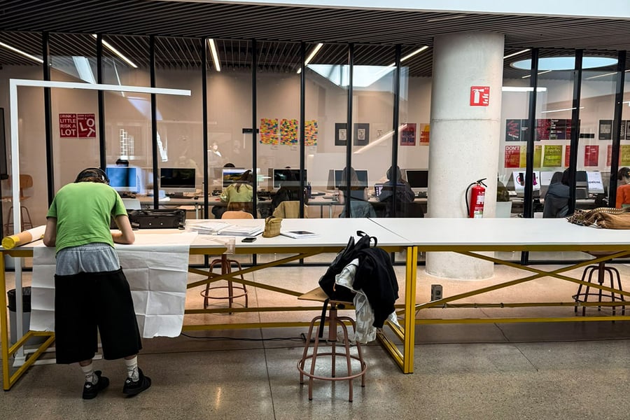 Eine Person arbeitet an einem langen, gelben Zeichentisch im Vordergrund, während durch eine raumhohe Glaswand im Hintergrund ein Computerraum mit arbeitenden Studierenden sichtbar ist. An den Wänden des Raums hängen bunte Grafikdesign-Poster.