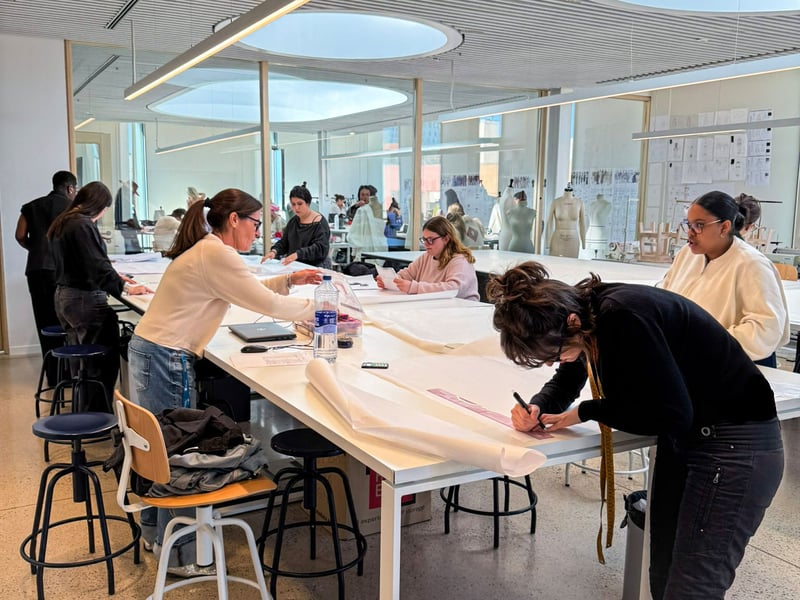 Eine Gruppe von Modedesign-Studierenden arbeitet an großen, weißen Tischen in einem hellen Atelier der LCI Barcelona. Sie zeichnen Schnittmuster auf lange Papierrollen und tauschen sich aus, während im Hintergrund Schneiderpuppen und Entwürfe an den Wänden zu sehen sind.