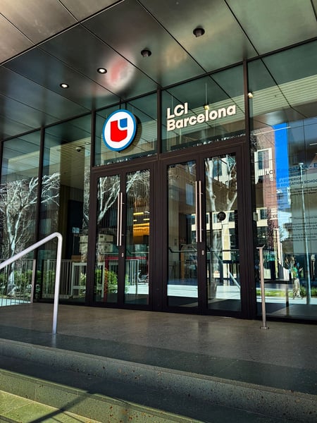 Der Haupteingang der LCI Barcelona mit einer modernen Glasfront und einer dunklen Metalltür. Über dem Eingang prangt das blau-rote Logo der Designschule sowie der Schriftzug ‚LCI Barcelona‘, während sich in der Verglasung die umliegenden Gebäude und Bäume spiegeln.