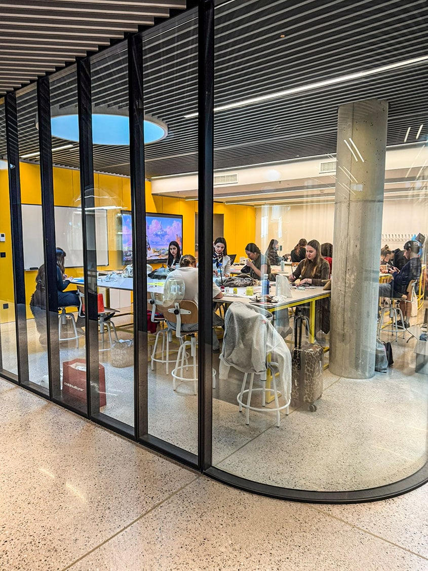Blick durch eine geschwungene Glaswand in einen modernen Seminarraum der LCI Barcelona. Eine Gruppe von Studierenden sitzt an einem großen Gemeinschaftstisch und arbeitet konzentriert, während im Hintergrund eine leuchtend gelbe Wand und ein großer Monitor zu sehen sind.