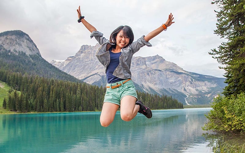 Entdecke Kanada mit AIFS: Frau springt am smaragdgrünen See vor majestätischen Bergen.