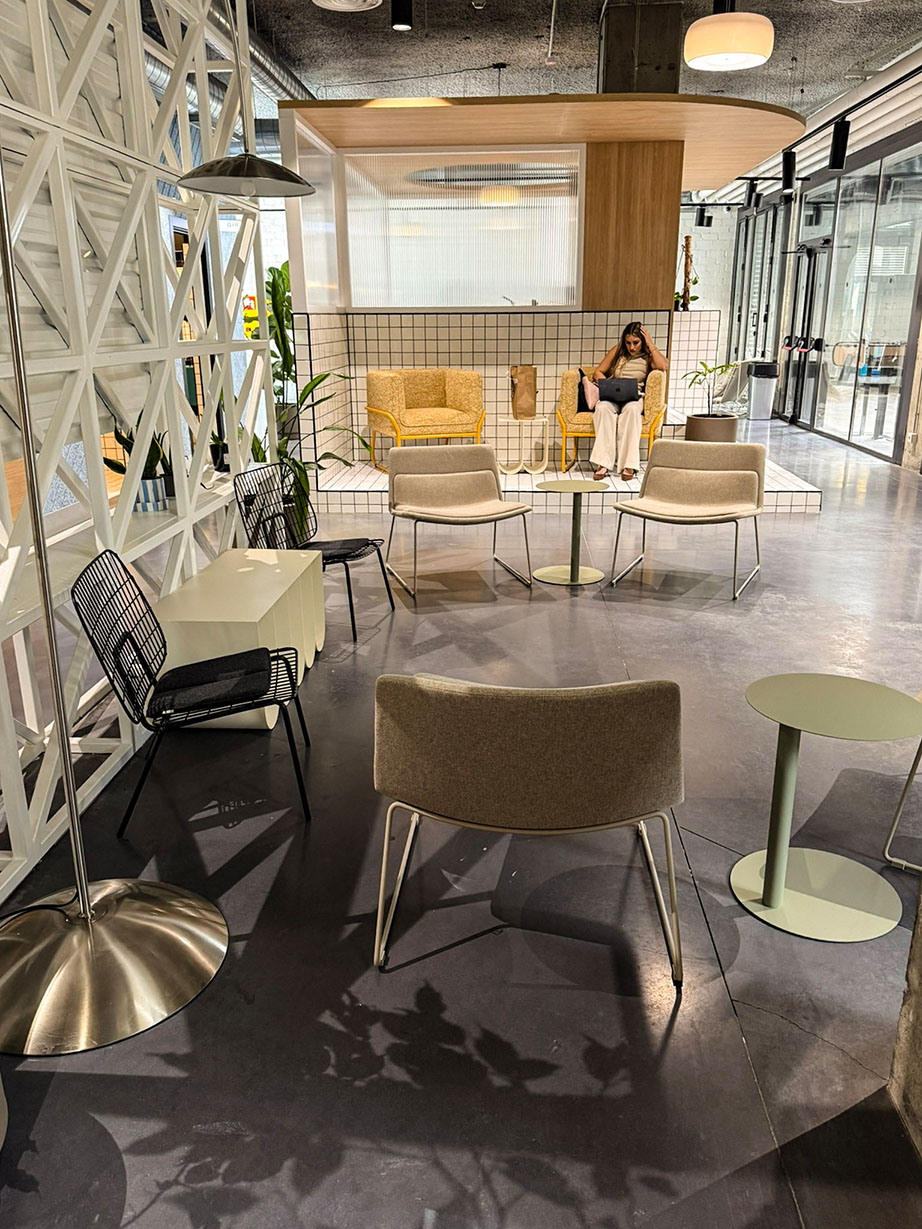 Ein stilvoller Gemeinschaftsbereich in der Unterkunft mit einer Mischung aus modernen Designer-Stühlen und kleinen Beistelltischen auf einem dunklen Boden. Im Hintergrund arbeitet eine Person in einer gelben Sitznische, die mit weißen Kacheln und einer markanten Holzüberdachung gestaltet ist.