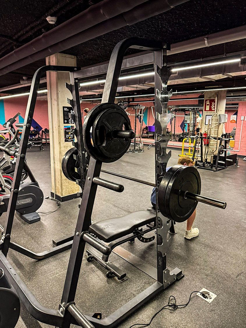 Ein modern ausgestattetes Fitnessstudio mit Fokus auf eine Hantelbank und ein Power-Rack mit schweren Gewichtsscheiben im Vordergrund. Der Raum hat einen dunklen Gummiboden, Sichtbetonsäulen und rosafarbene Akzentwände, während im Hintergrund weitere Trainingsgeräte zu sehen sind.