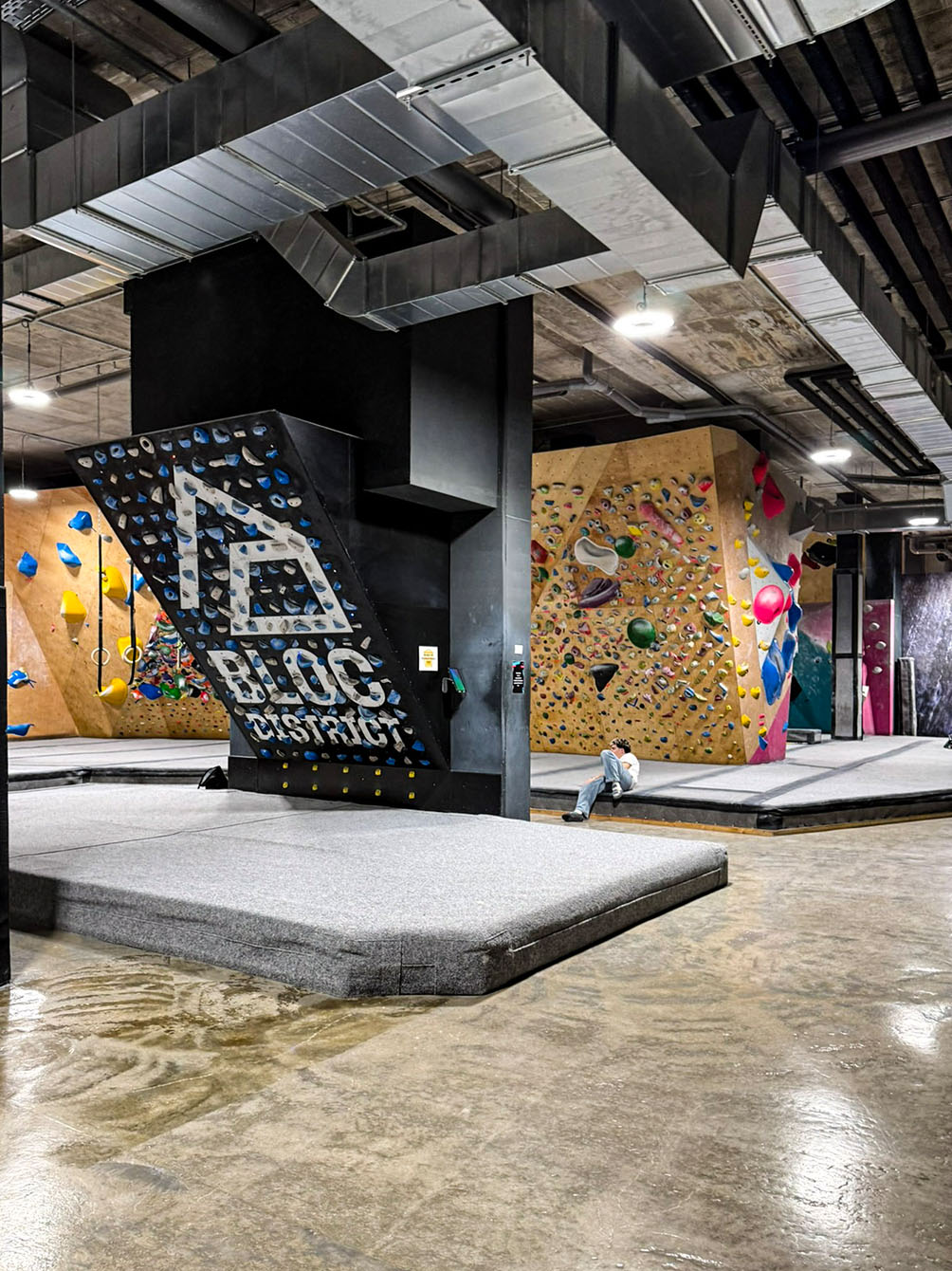Eine weitläufige Innenansicht der Boulderhalle ‚Bloc District‘ mit mehreren Kletterwänden, die mit bunten Griffen bestückt sind. Im Zentrum steht ein schwarzer Pfeiler mit dem Logo der Halle, während im Hintergrund eine Person auf den grauen Sicherheitsmatten Pause macht.